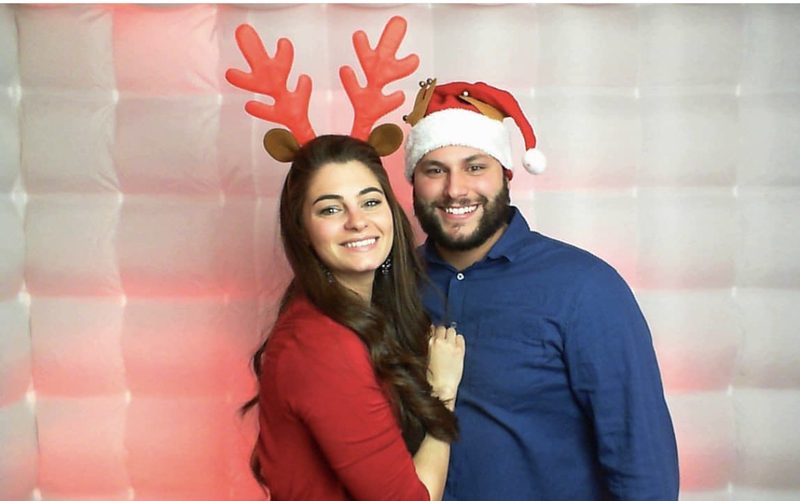 A couple in reindeer antlers and a Santa hat in front of the lit booth backdrop