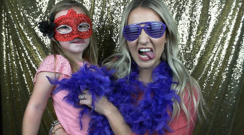 A guest holding her daughter, both in sequin masks, shutter shades and feather boas against the gold backdrop