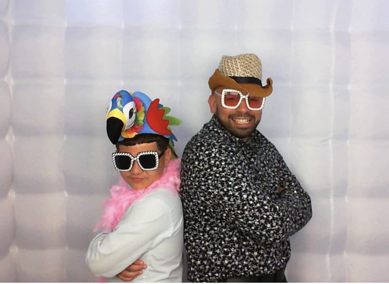A child in a parrot hat and pink feather boa standing back to back with a guest in a cowboy hat and jeweled glasses