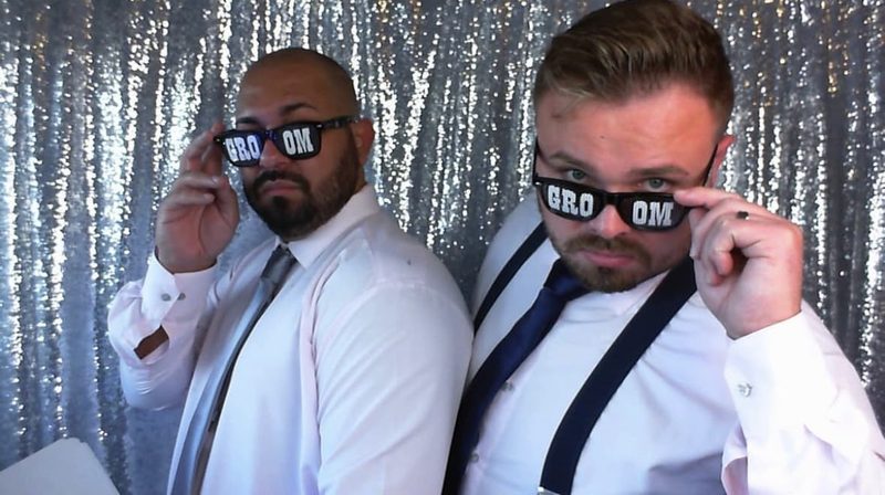 Two grooms in shirts and ties tilting novelty “GROOM” glasses, against a silver sequin backdrop