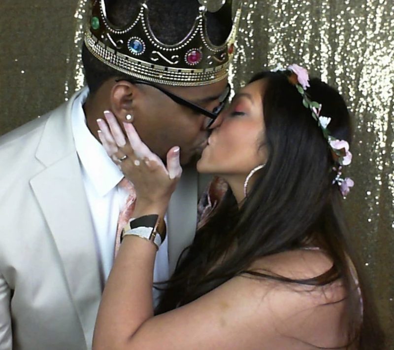 A couple sharing a kiss, one wearing a gold crown and the other a flower crown, against the gold sequin backdrop